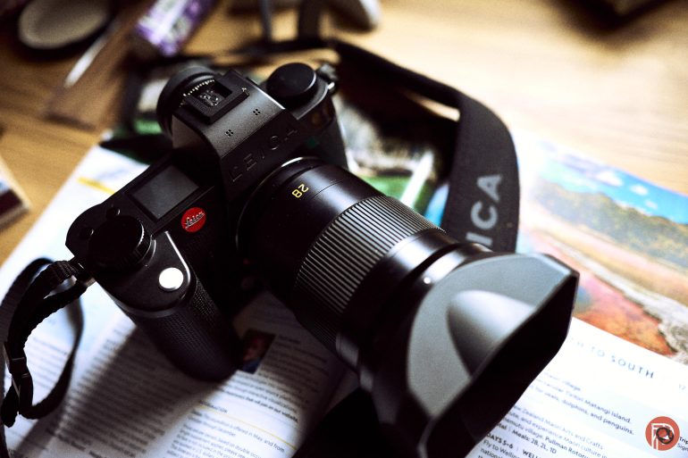 A black Leica camera with a zoom lens sits on an open magazine on a wooden surface.
