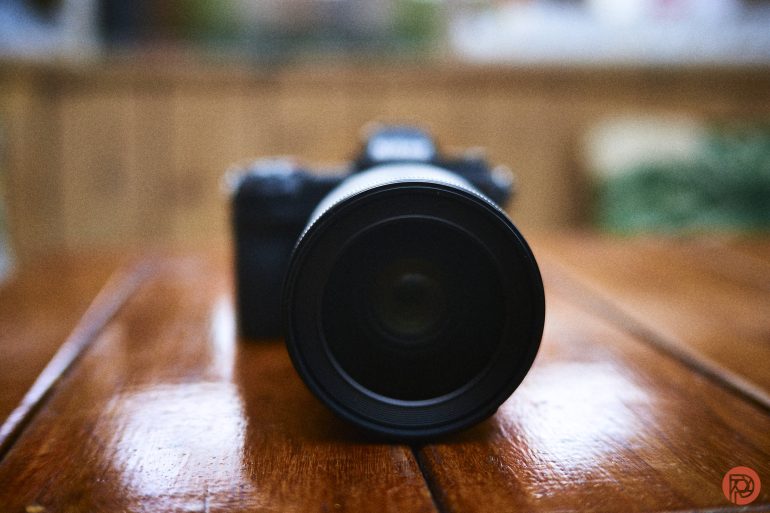 A black camera with a large lens sits on a wooden surface, photographed from the front.