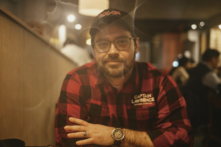 Man in glasses and a cap, wearing a red plaid shirt, sitting with arms crossed in a dimly lit room.