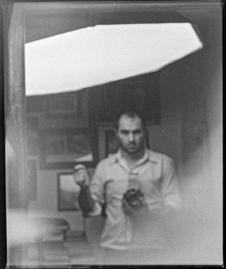 A man captures a vintage mirror selfie, holding his DIY camera and gazing intently into the reflection, with blurred surroundings adding to the timeless aura.
