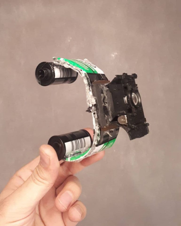 A hand holds a DIY pinhole camera crafted from film canisters and cardboard.