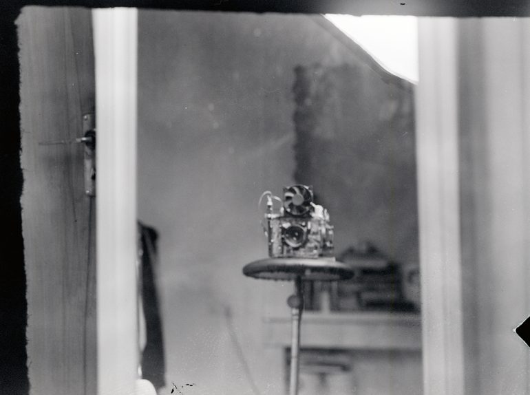 A dimly lit room reveals a blurry vintage DIY camera perched on a tripod, as seen through a doorway.