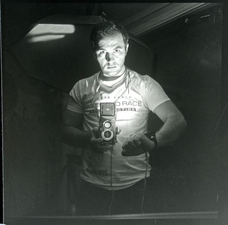 In a dimly lit room, a man holds a DIY vintage camera, wearing a T-shirt with text, and gazes intently at the lens.