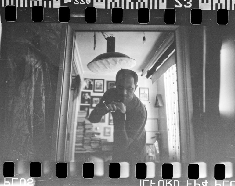 A black and white film photo captures a man using a DIY camera for a mirror selfie in a room adorned with photos on the wall.