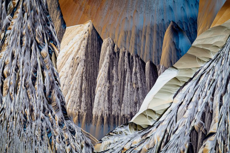 An abstract image resembling a surreal landscape with jagged, mountain-like formations. The layered texture, with shades of gray, brown, and blue, suggests a captivating blend of natural and artistic elements. Image by Rob Blanken.