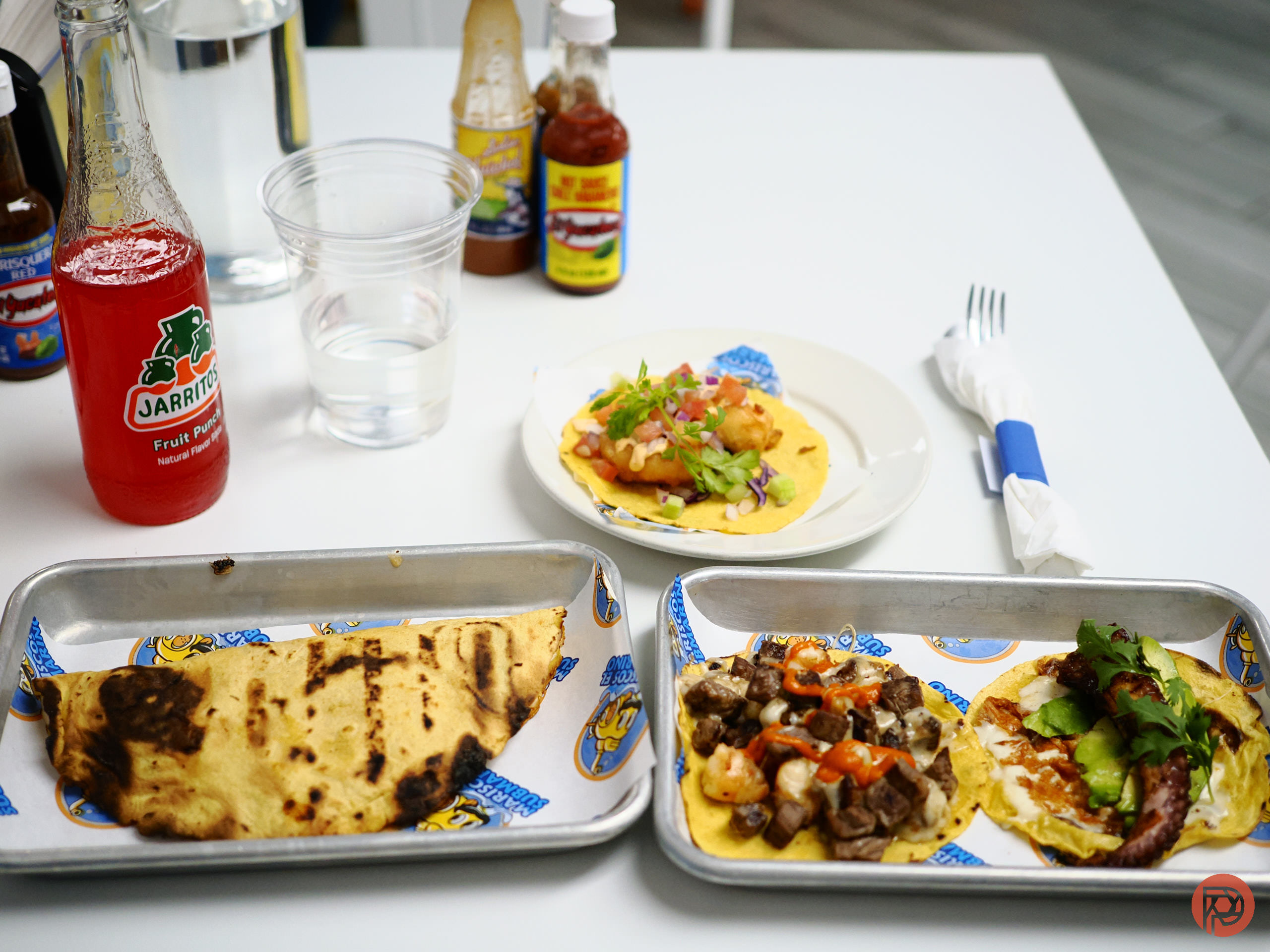 Three plates with various tacos, Jarritos soda, hot sauce bottles, and a glass of water on a white table.