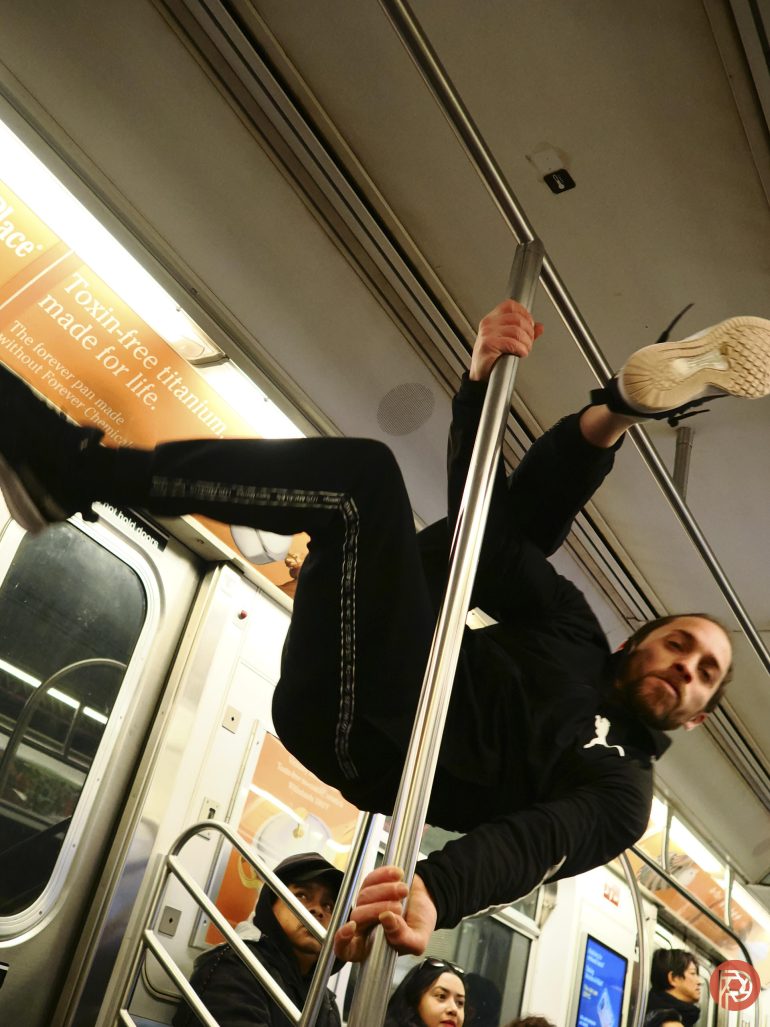 A man in athletic wear does a pole trick on a subway, holding on sideways with one leg extended up.