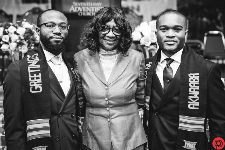 Three people in formal attire pose together, two wearing stoles with GREETINGS and AKWAABA.