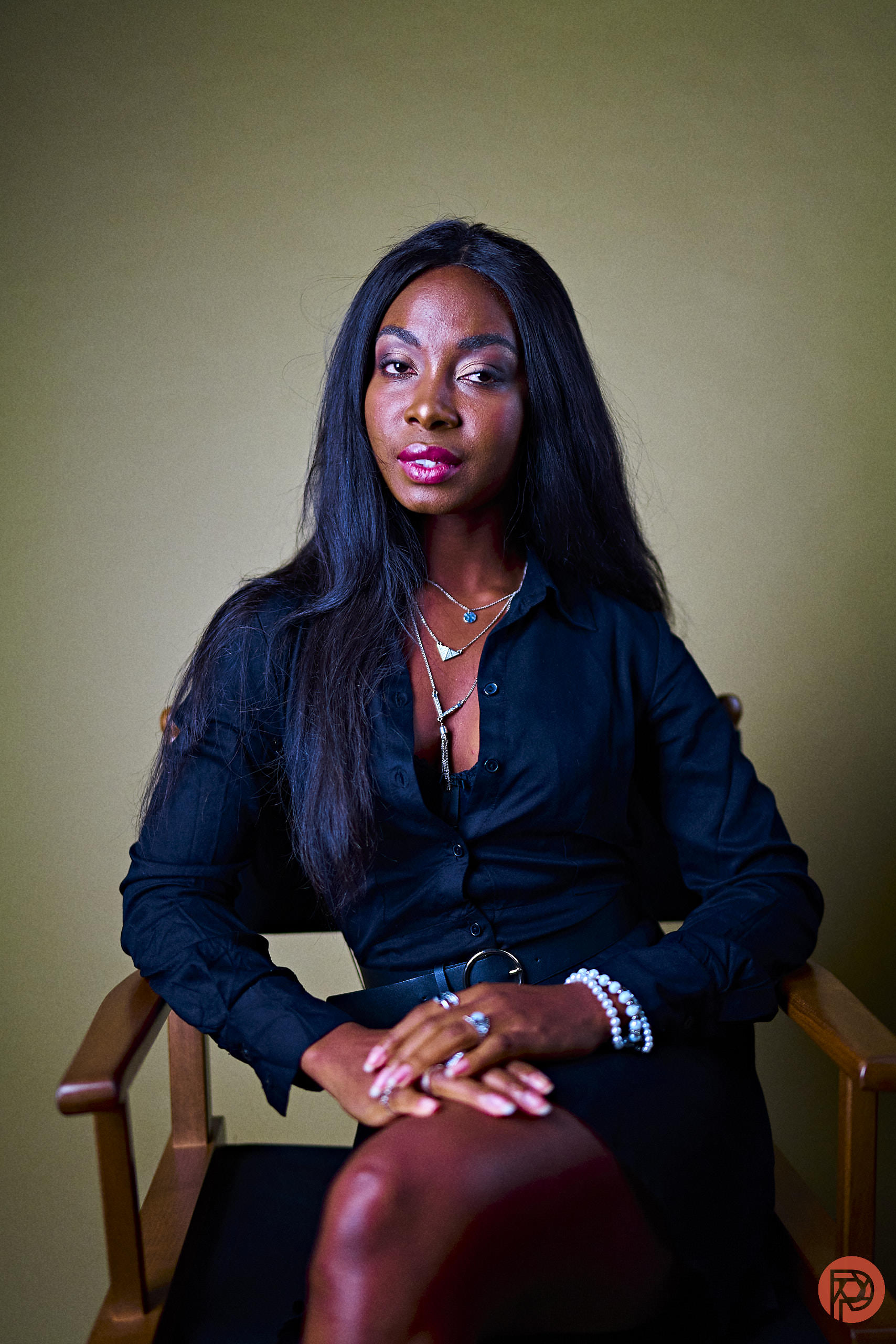 Woman in a black dress sits on a chair against a beige background, with hands folded on her lap.