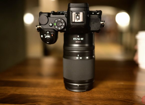 A Nikon camera with a zoom lens sits upright on a wooden table, blurred background and hand holding a cup.