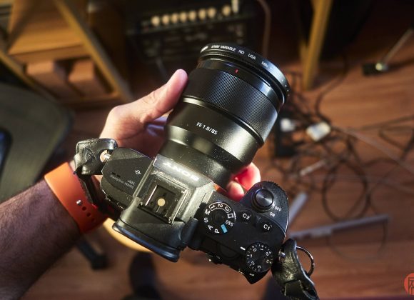 A person holding a Sony camera with an 85mm lens over a cluttered workspace with cables on the floor.