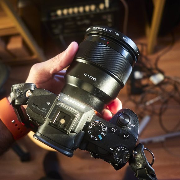 A person holding a Sony camera with an 85mm lens over a cluttered workspace with cables on the floor.