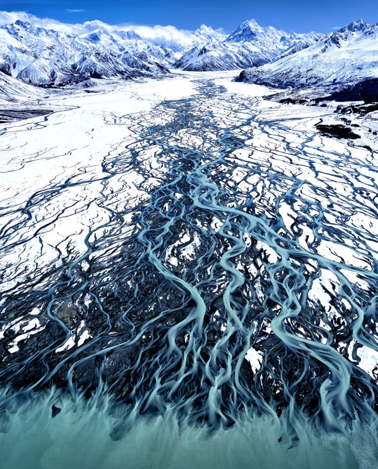 Aerial view of braided river patterns flowing through snow-covered mountains under a clear blue sky.