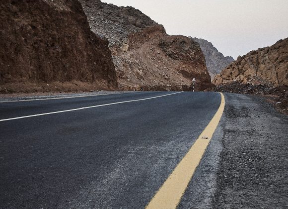 A winding mountain road with a yellow line on the right, flanked by rocky hills under a clear sky.