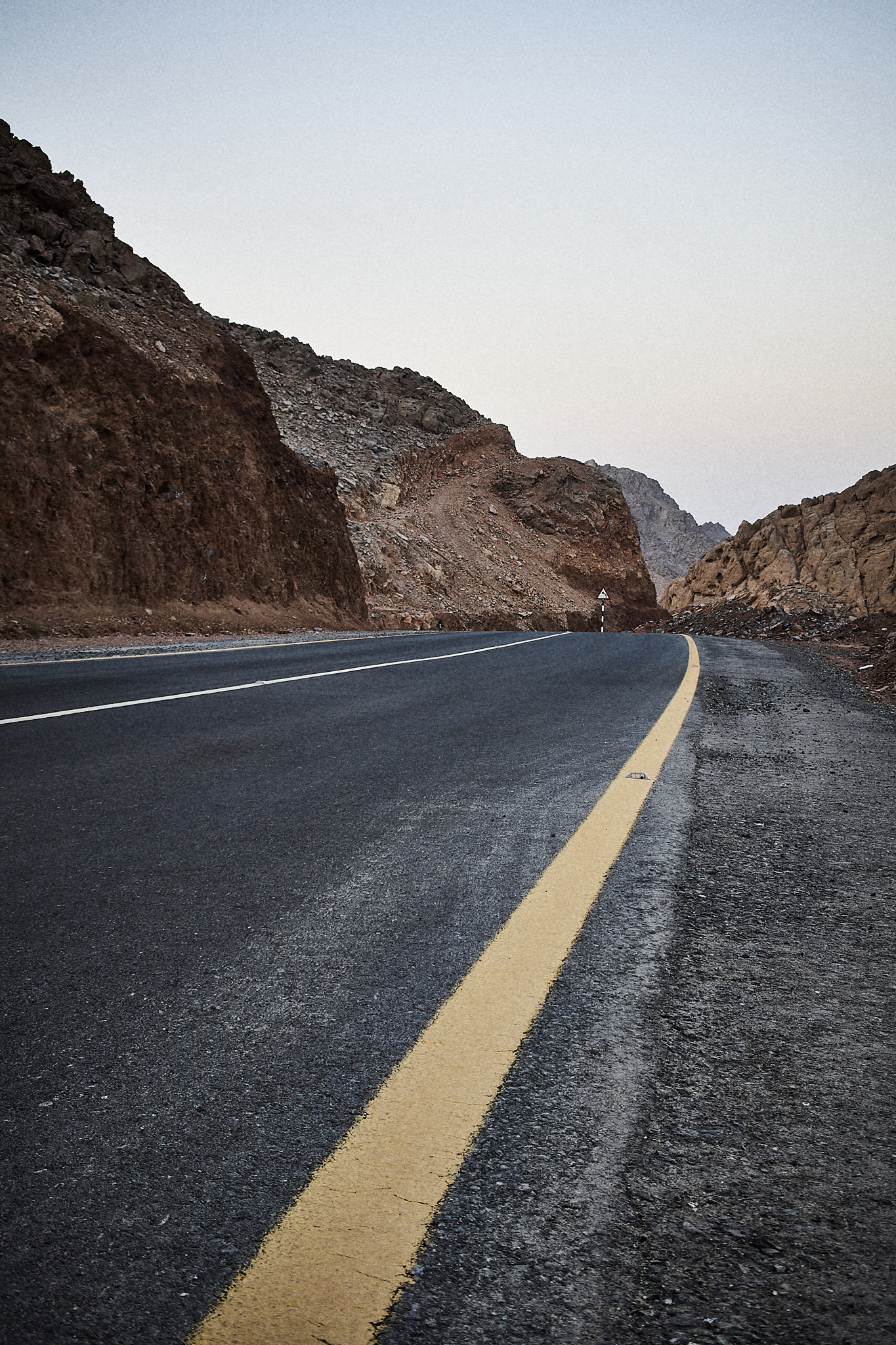 A winding mountain road with a yellow line on the right, flanked by rocky hills under a clear sky.