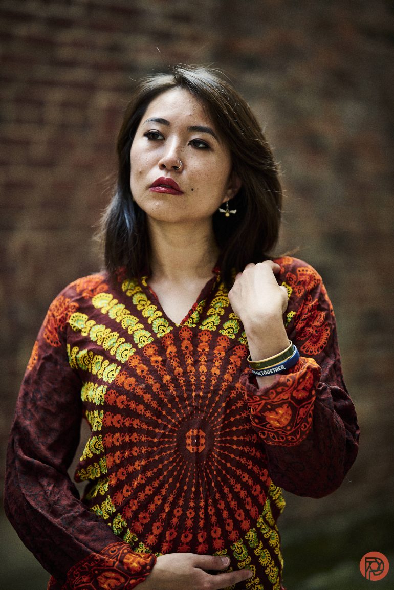 Person in a red and yellow patterned top stands against a blurred brick wall, looking slightly to the side.