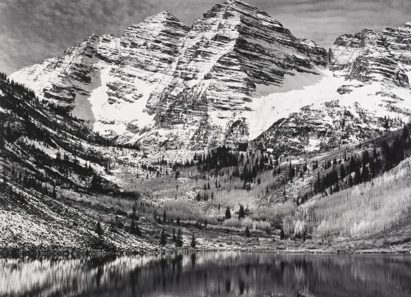Snow-covered mountain peaks reflected in a calm lake, with trees scattered on the slopes below.
