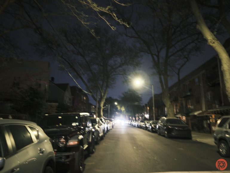 A blurry night view of a residential street lined with parked cars and leafless trees under streetlights.