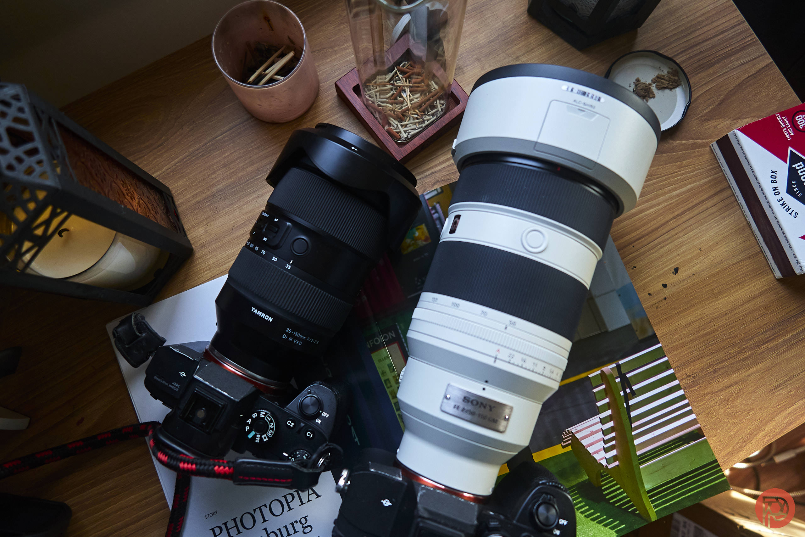 Two Sony cameras with large lenses rest on a wooden desk surrounded by books and small containers.