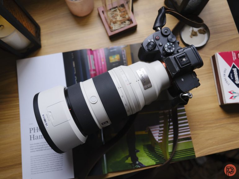 A DSLR camera with a large white telephoto lens rests on an open magazine on a wooden table.