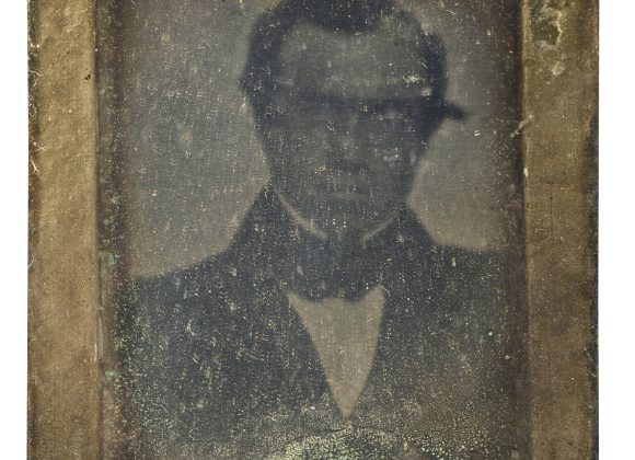 An old, faded daguerreotype portrait of a man in formal attire with a bow tie.