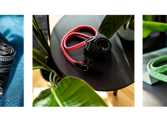 Three vintage cameras with colorful braided straps: yellow on magazine, red on table, green among plants.