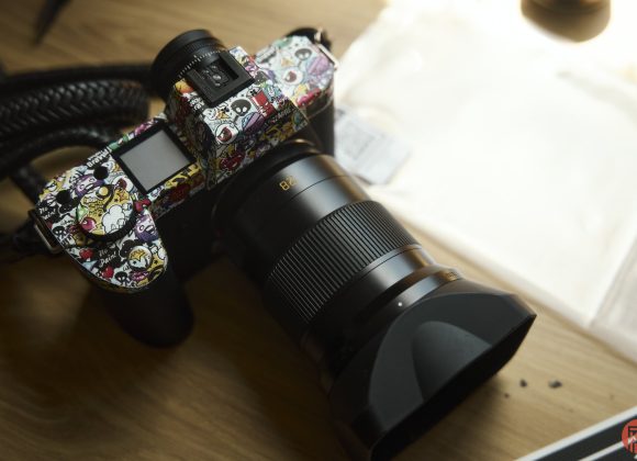 A camera with a colorful, cartoon-themed skin sits on a wooden table next to a white envelope.