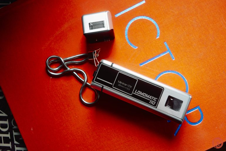 A vintage silver Lomomatic 110 camera with flash on an orange surface with blue lettering.