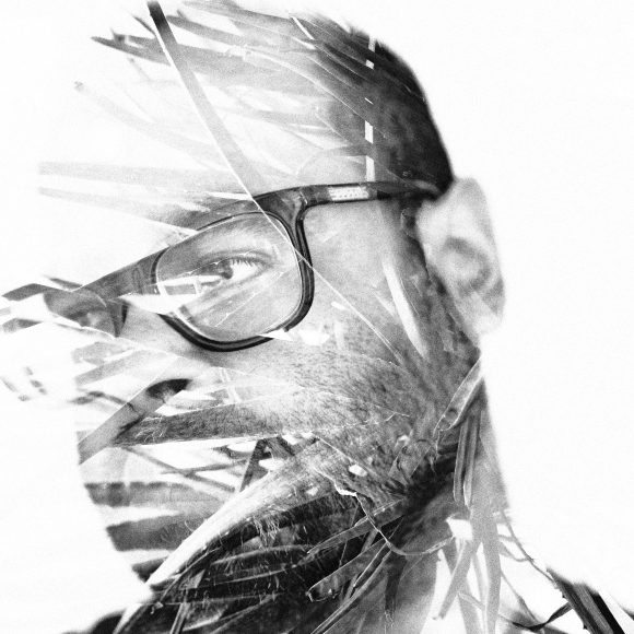 Black and white double exposure of a man wearing glasses overlaid with abstract palm leaves.