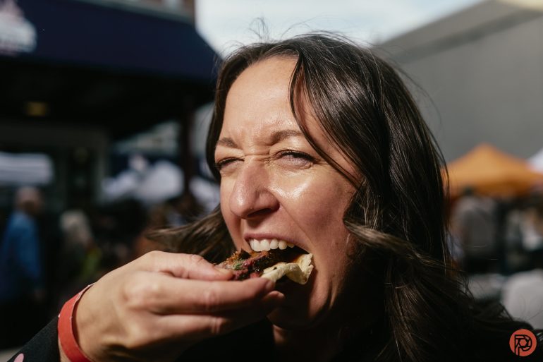 A woman smiling and eating food outdoors at an event, with a crowd and tents in the background.
