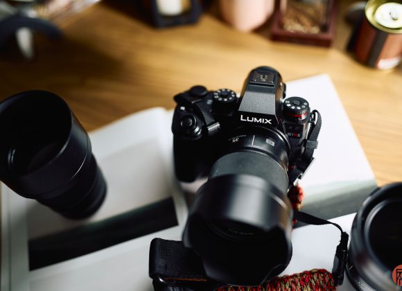 A Lumix camera with lens sits on an open book, surrounded by camera equipment on a wooden table.