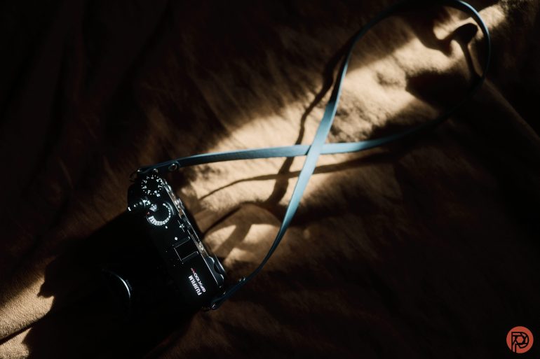 A camera with a blue strap lies on a brown fabric, illuminated by a beam of sunlight.