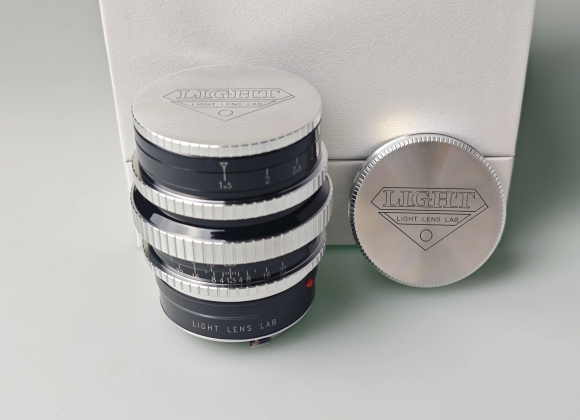 A silver and black camera lens with Light Lens Lab branding, next to a matching lens cap and white box.