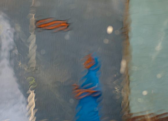 A person in a blue outfit is walking past a blue wall, seen through a rain-blurred window.