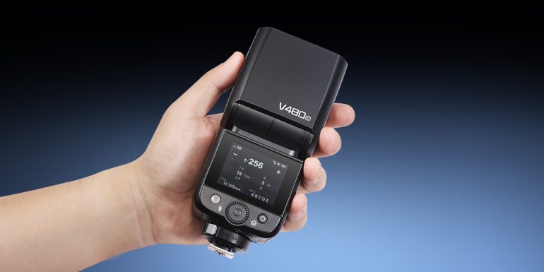 A hand holding a Godox V490 flash with a digital display and control buttons visible.