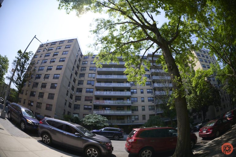 Wide-angle view of a tall apartment building with parked cars and leafy trees lining the street.