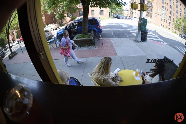 View from a cafe window of people sitting outside at a yellow table and a woman walking on the sidewalk.