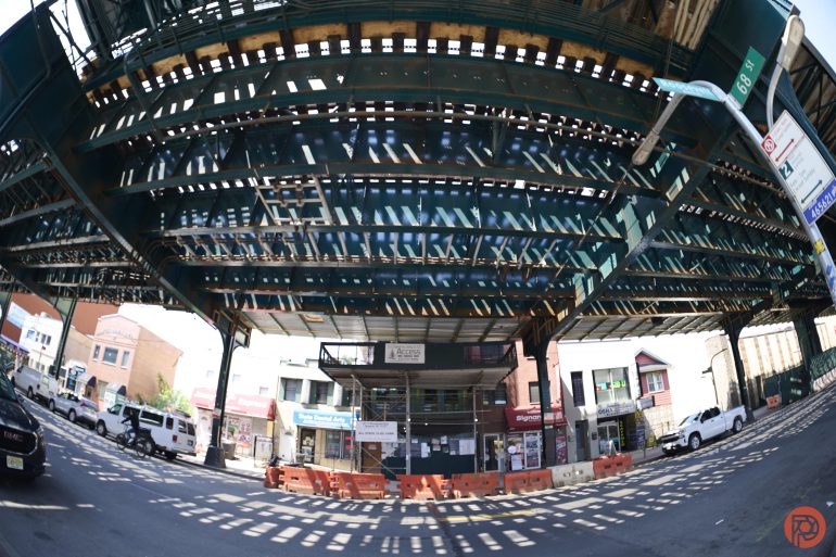 Wide view of an elevated train track casting shadows over a city street with construction barriers below.