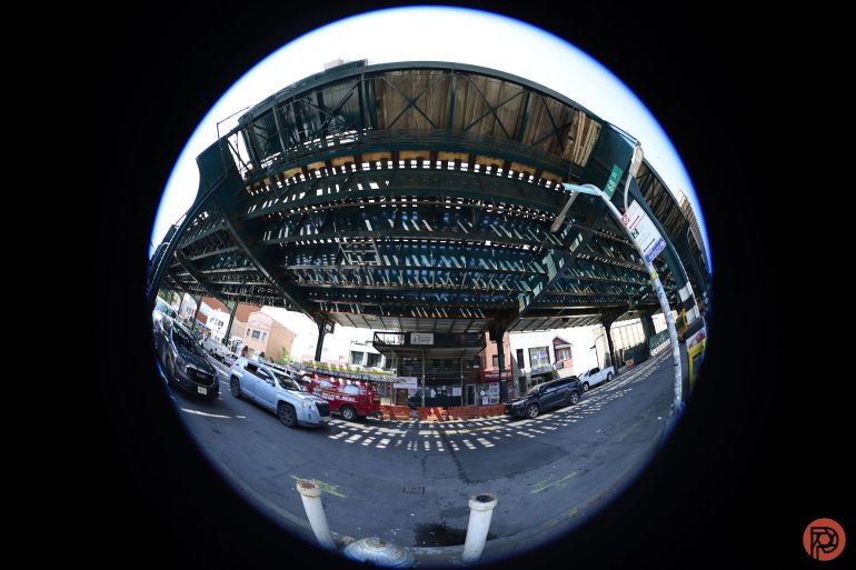 Fisheye view of an elevated train track over a busy urban street with cars and buildings below.