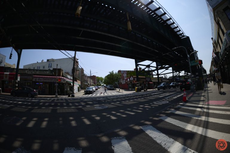 A city street with elevated train tracks casting shadows across the road and buildings.