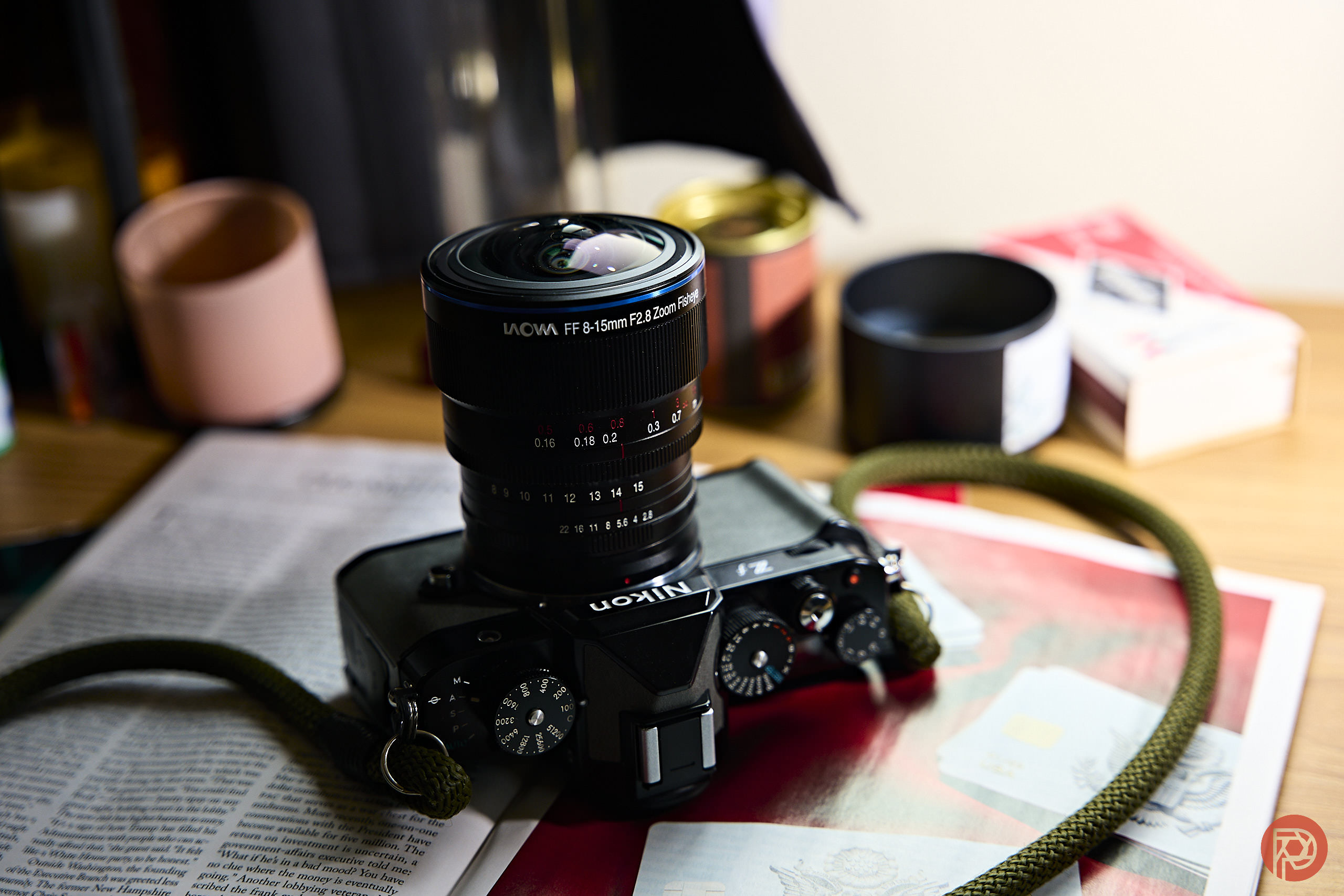A Nikon camera with a LAOWA lens rests on an open newspaper, surrounded by cups and small boxes.
