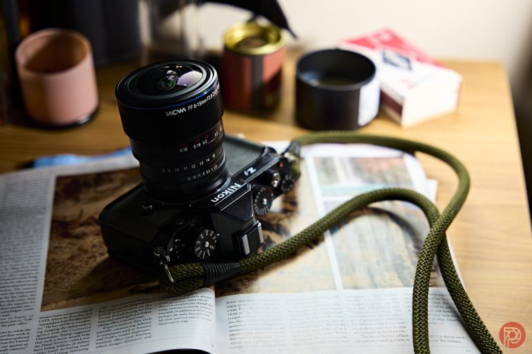 A camera with a green strap rests on an open magazine on a wooden table with cups in the background.