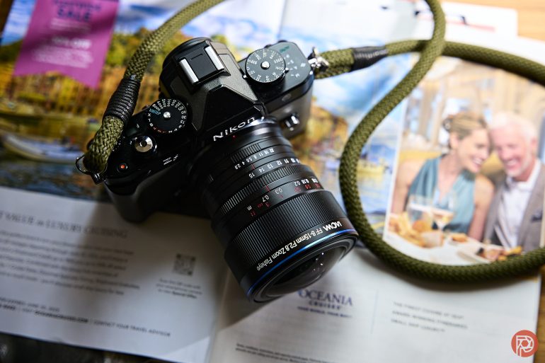A Nikon camera with a green strap rests on open magazines featuring travel ads and a couple dining.
