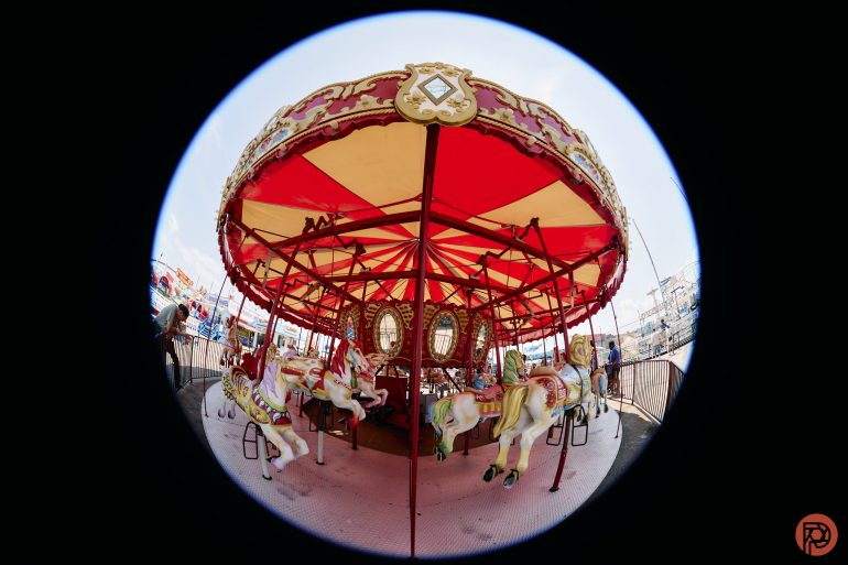 Fisheye view of an empty, vintage carousel with colorful horses under a red and yellow canopy.