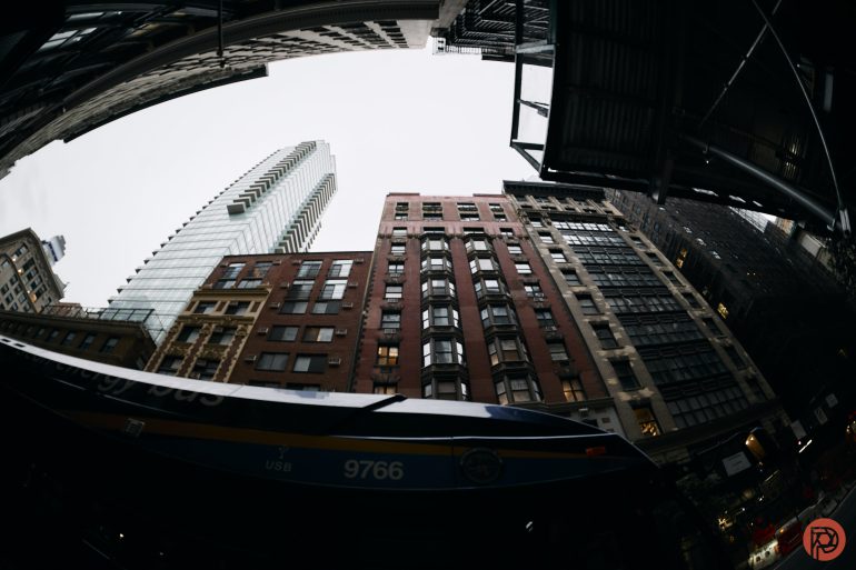 Wide-angle view of tall city buildings with a bus passing below on a cloudy day.
