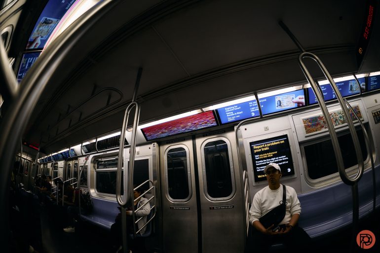 A person in a white shirt sits alone on a subway train with bright screens and empty seats.