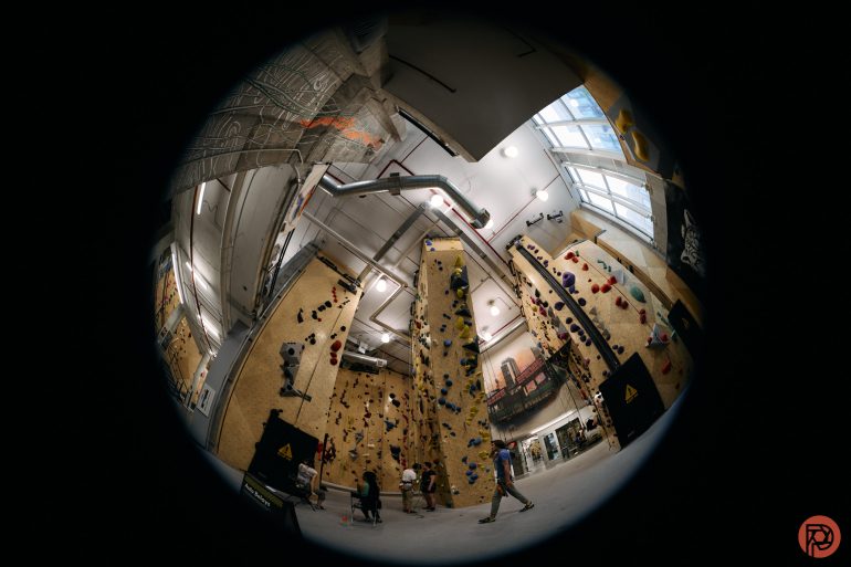 Fisheye view of an indoor rock climbing gym with people climbing tall walls and others watching below.