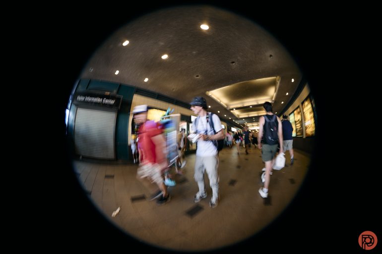 A fisheye lens photo of people walking in a busy indoor corridor with a closed shop on the left.