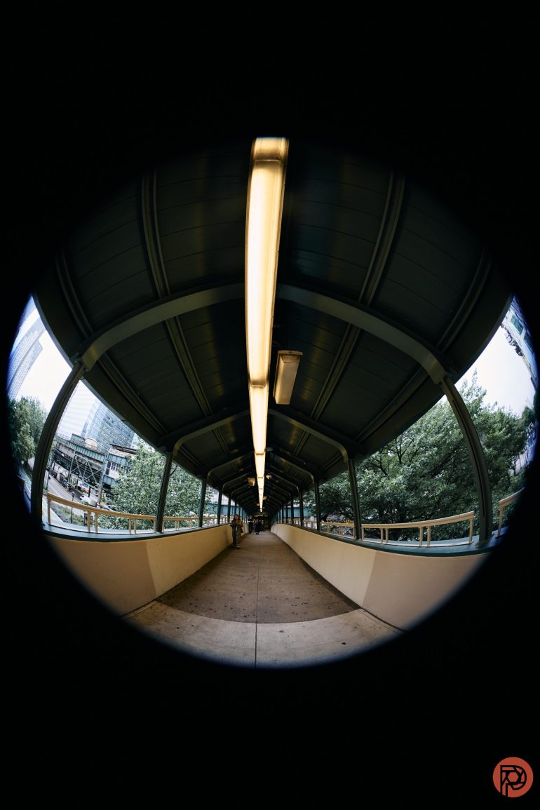A fisheye view of a covered pedestrian walkway with a lit ceiling and trees visible outside.