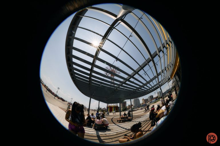 People sit under a large shaded structure at a sunny outdoor venue, seen through a fisheye lens.